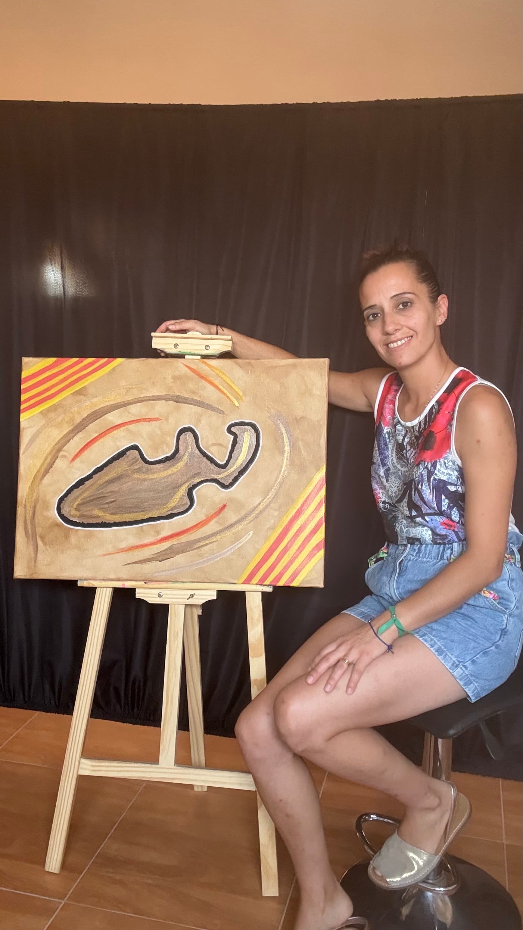 Mujer sonriente sentada junto a un caballete con una pintura abstracta de formas orgánicas marrones.